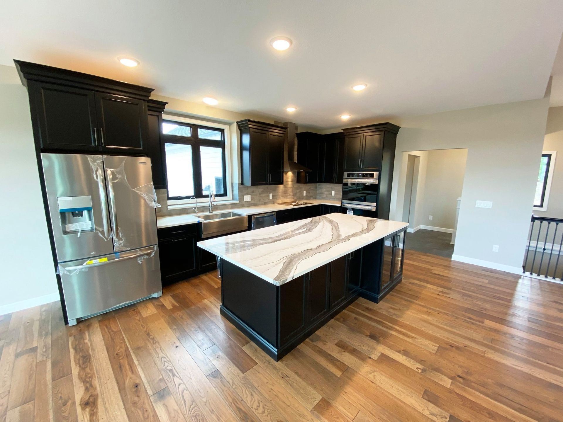 Modern kitchen with black cabinets, stainless steel fridge, marble island, and hardwood floors.