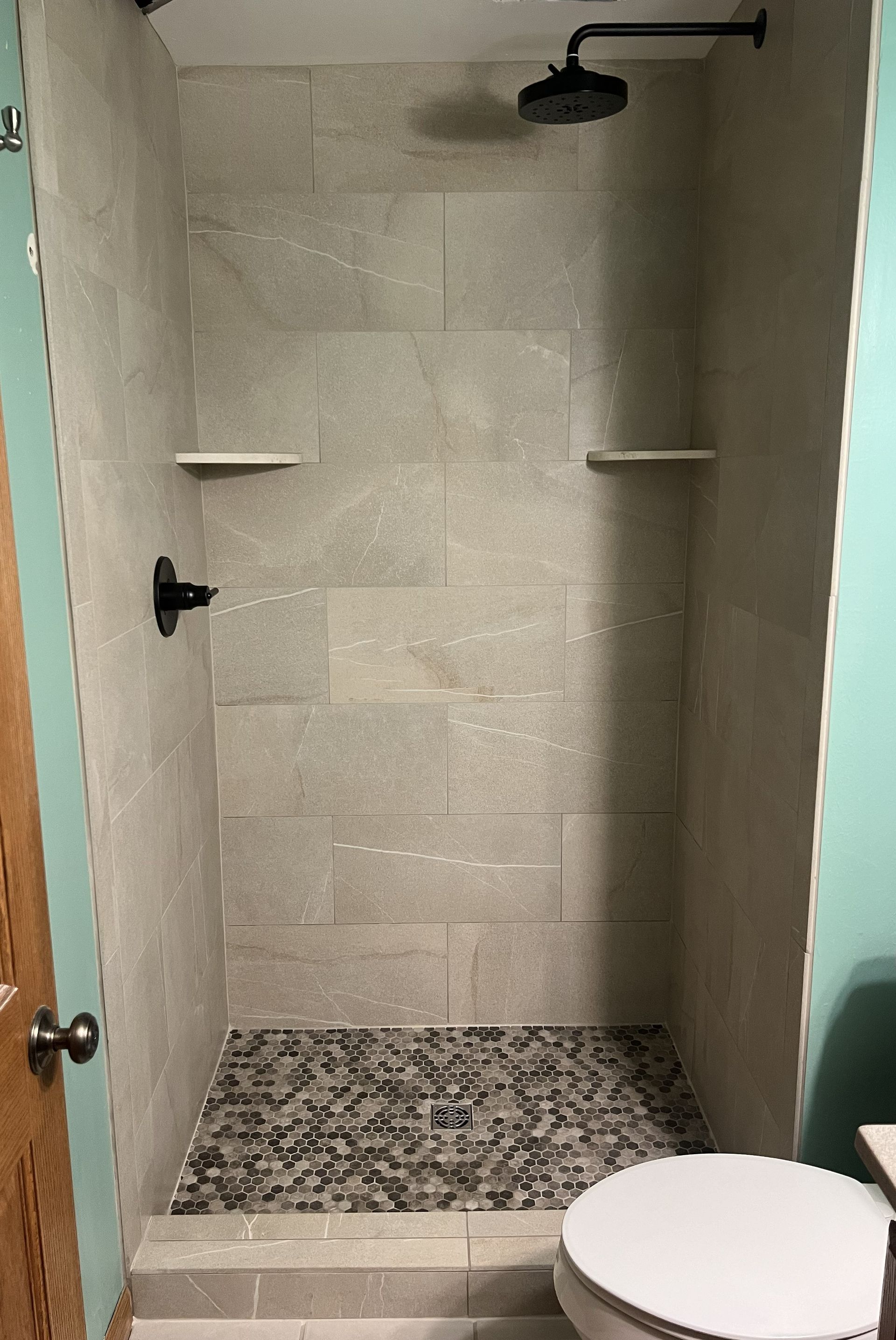 Empty tiled walk-in shower with black fixtures, corner shelves, and patterned mosaic floor.