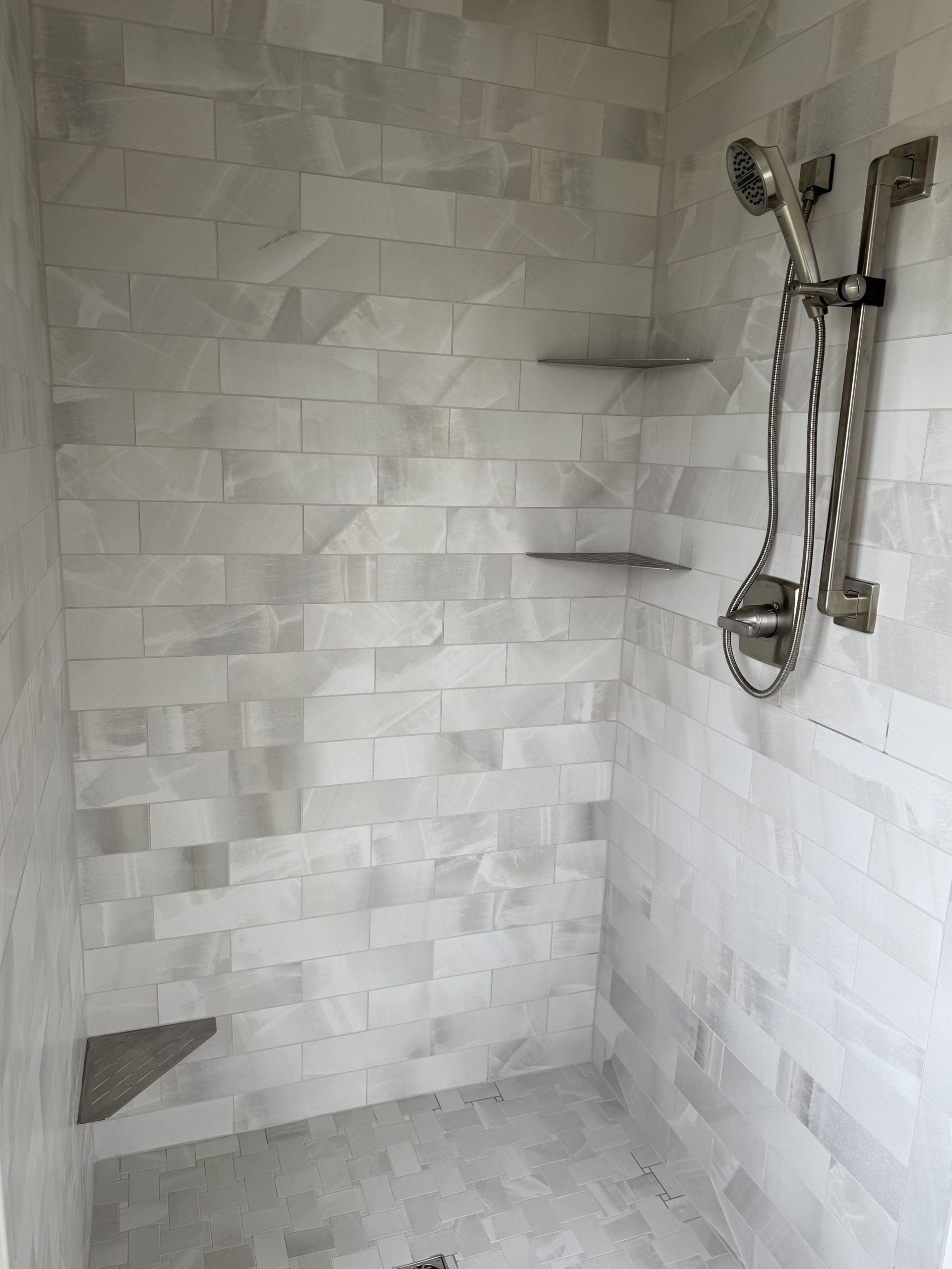 Tiled walk-in shower with showerhead, faucet controls, and small corner shelf in a gray bathroom