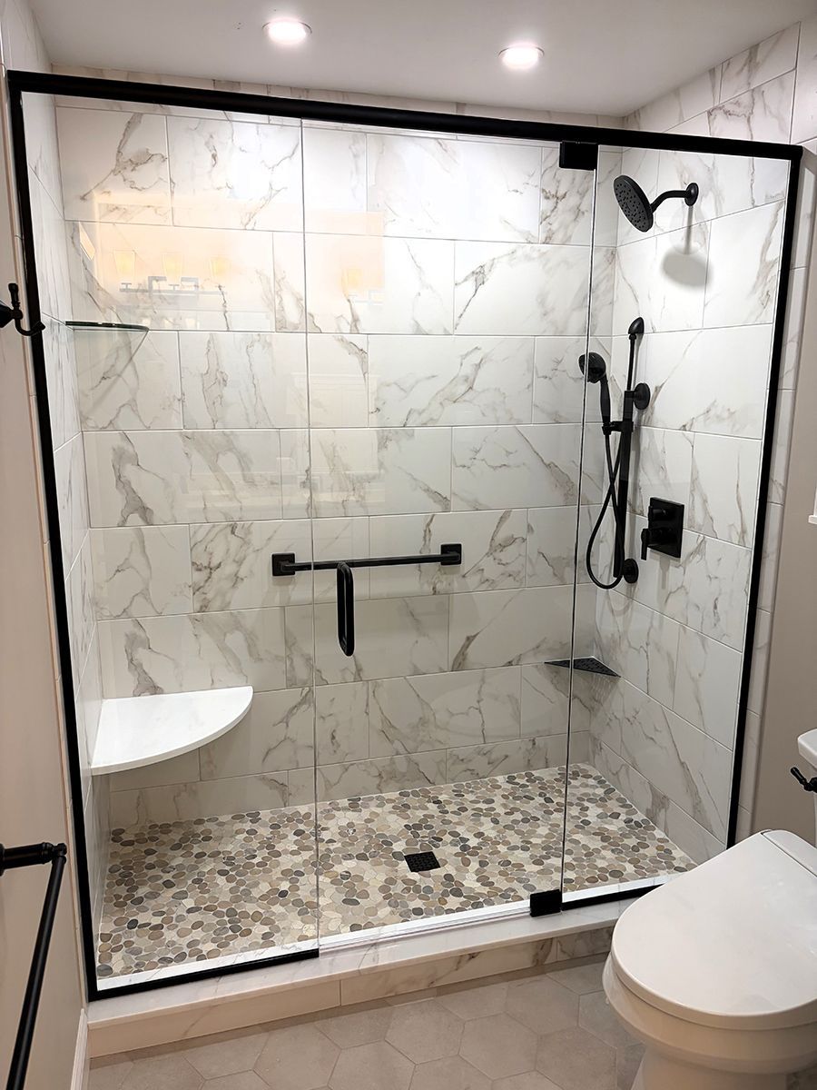 Accessible walk-in shower with marble tile, black grab bars, handheld showerhead, and toilet in foreground