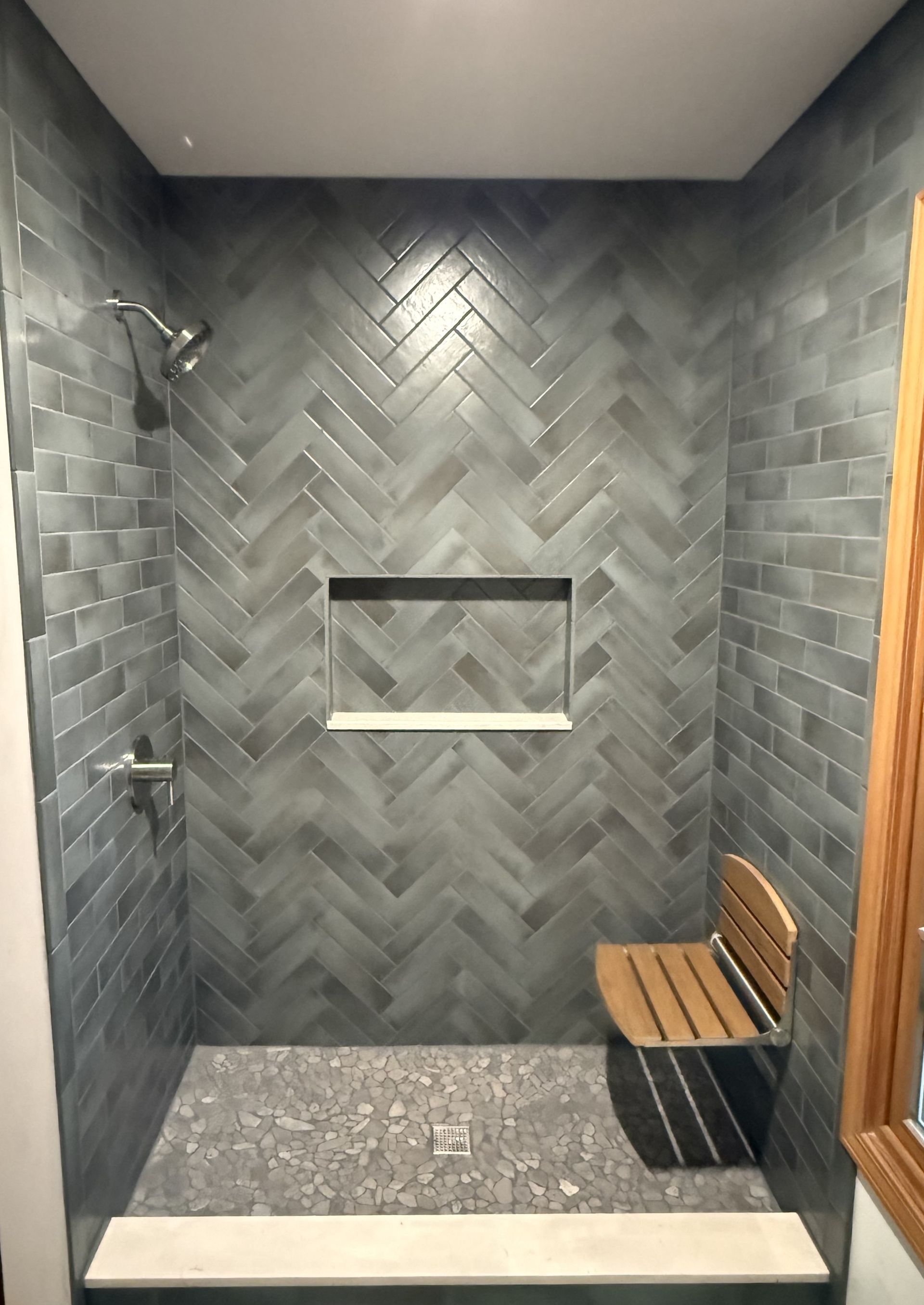 Modern gray-tiled shower stall with built-in niche, pebble floor, and wooden bench.