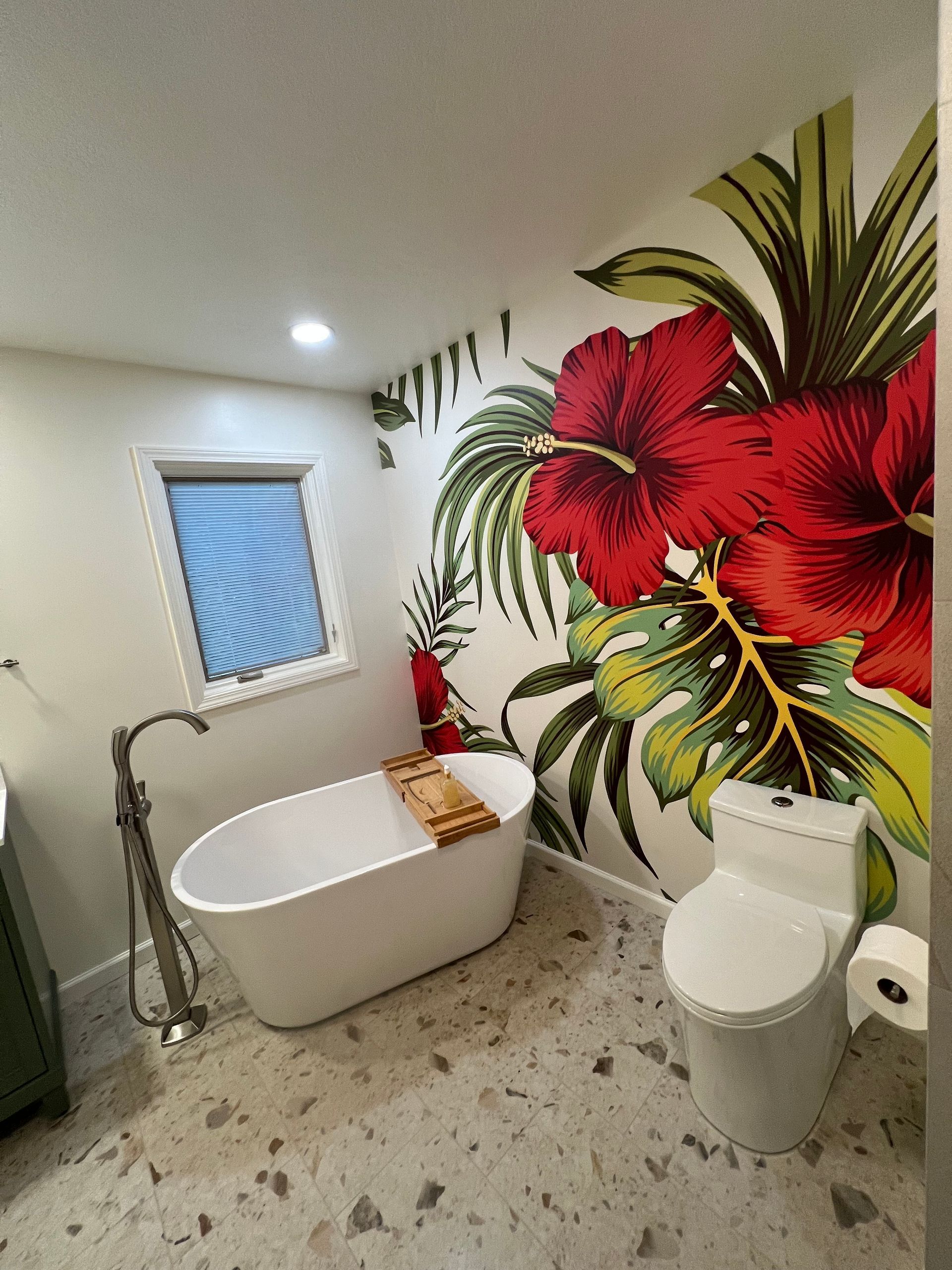 Modern bathroom with floral mural, freestanding tub, toilet, and window with blind