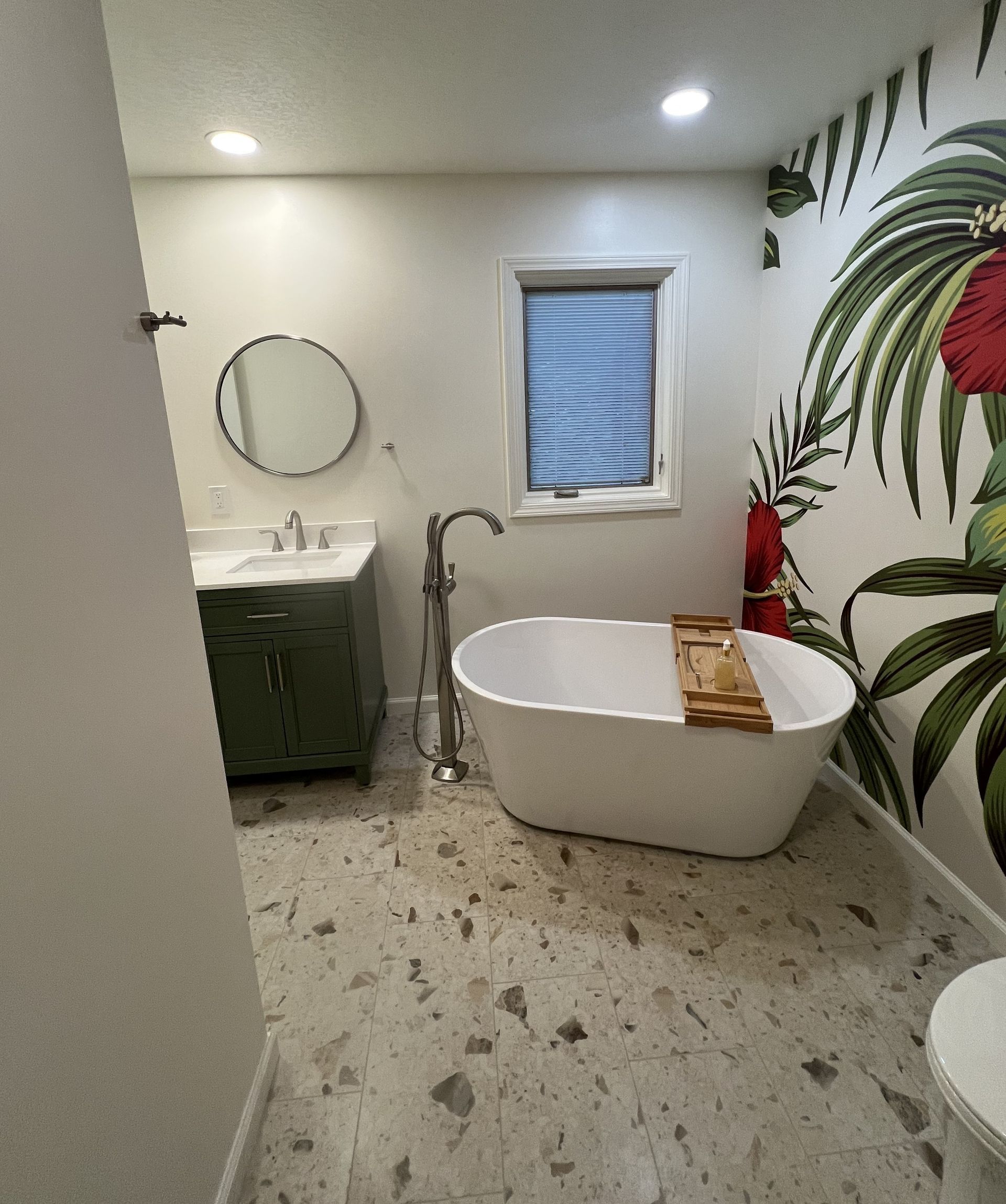 Modern bathroom with freestanding tub, green vanity, round mirror, window, and tropical wall mural