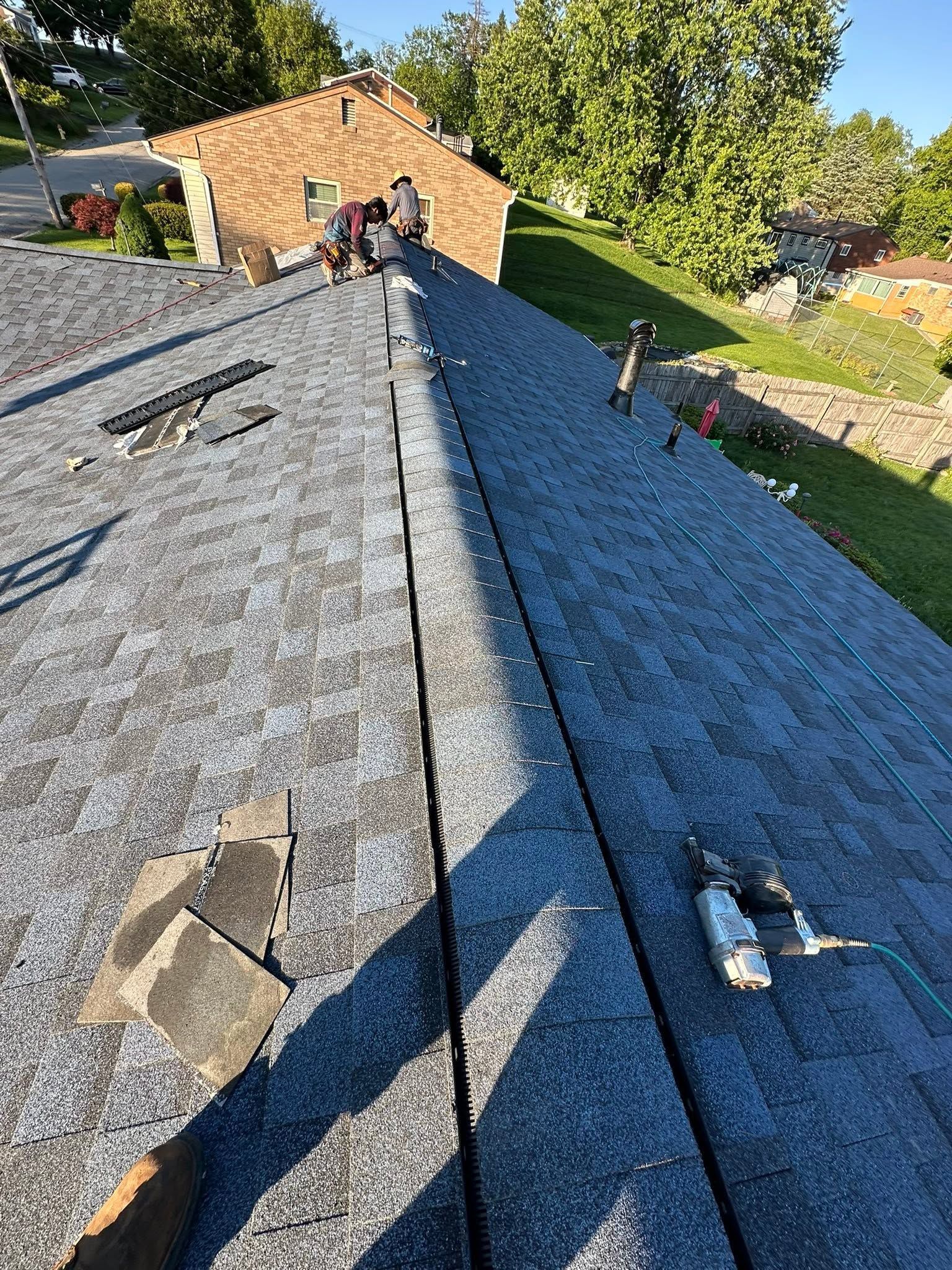 Two workers on a gray shingled roof beside a brick house, with tools and a ladder nearby.
