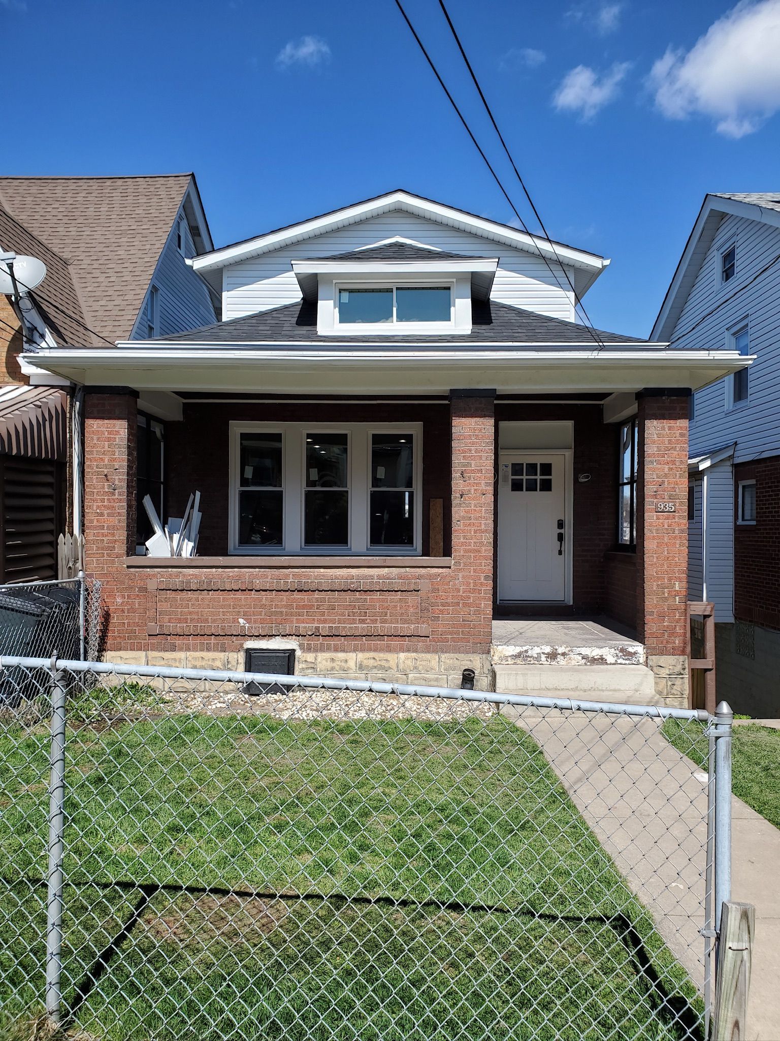 Small brick house with a front porch, white trim, and a chain-link fence under a clear blue sky.