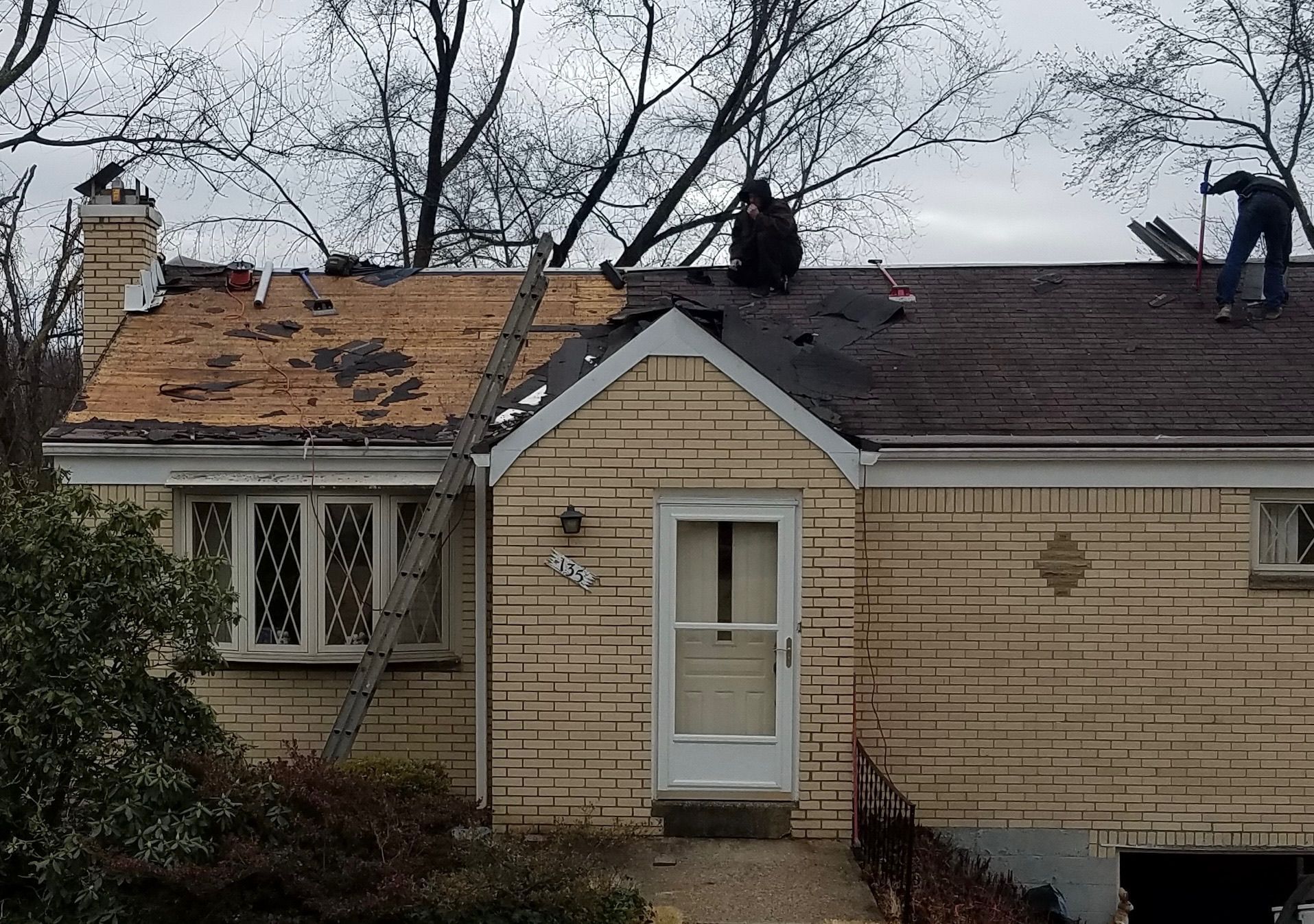 Workers repairing a house roof with exposed plywood and shingles on a cloudy day