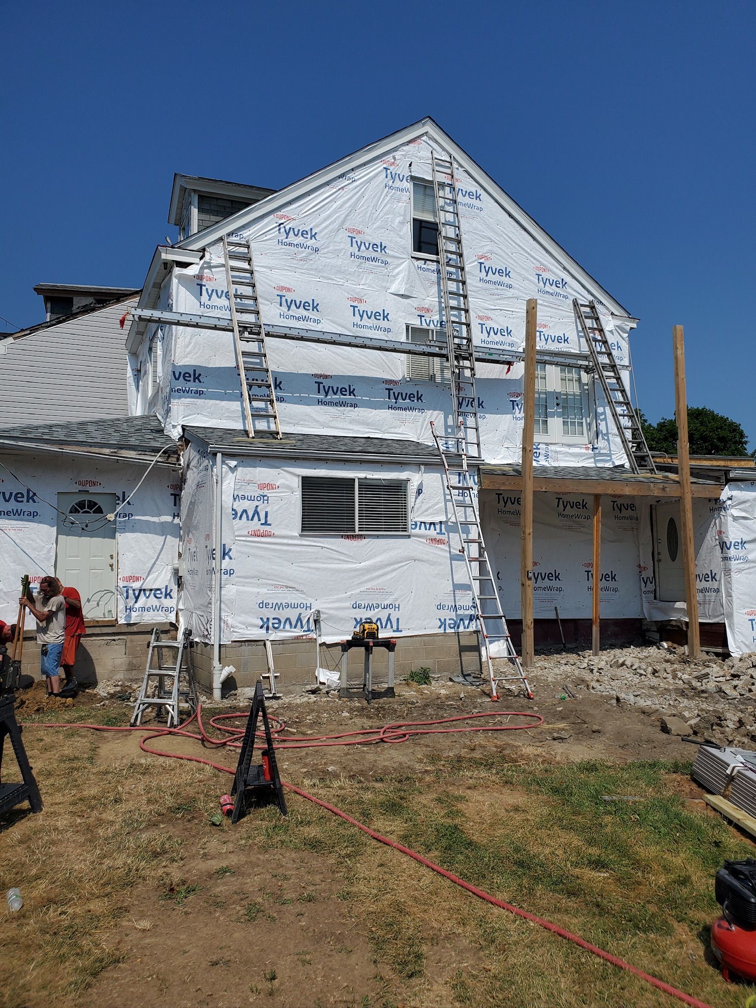White house with storm damage, peeling siding, and scattered debris on a muddy lot under clear blue sky