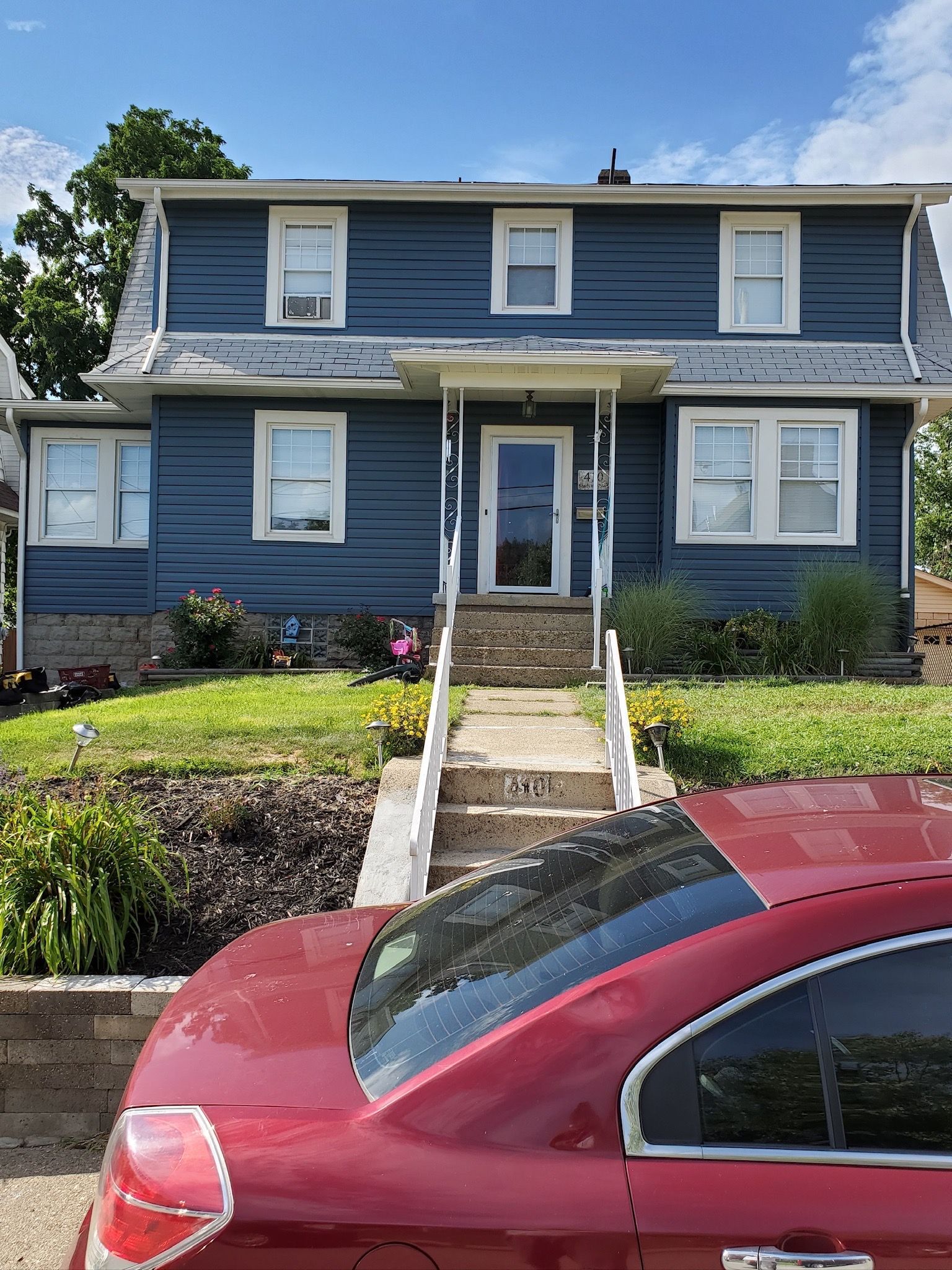 Blue two-story house with front steps, white trim, and a red car parked in front under a clear sky