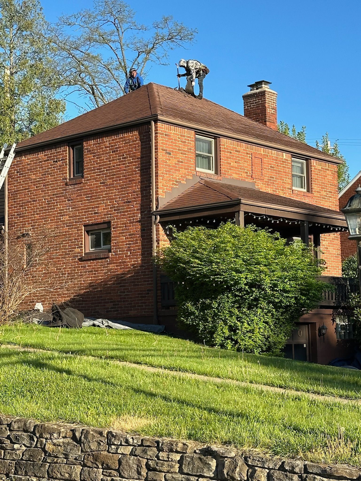 Red brick house with a porch on a sunny hillside, surrounded by green grass and trees.