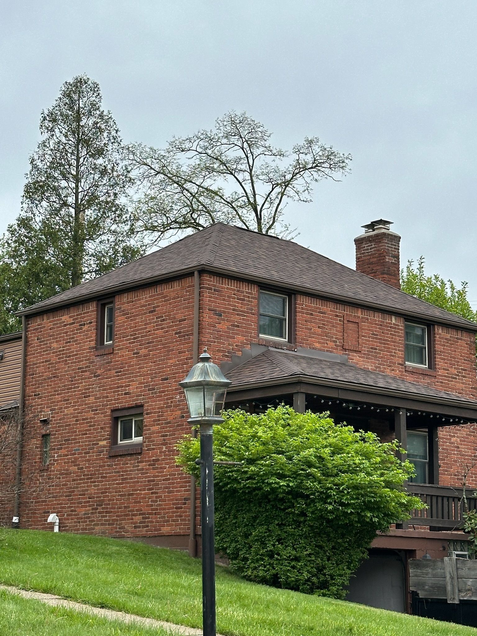 Brick two-story house with a tiled roof, chimney, and porch behind a lamp post and green shrub.