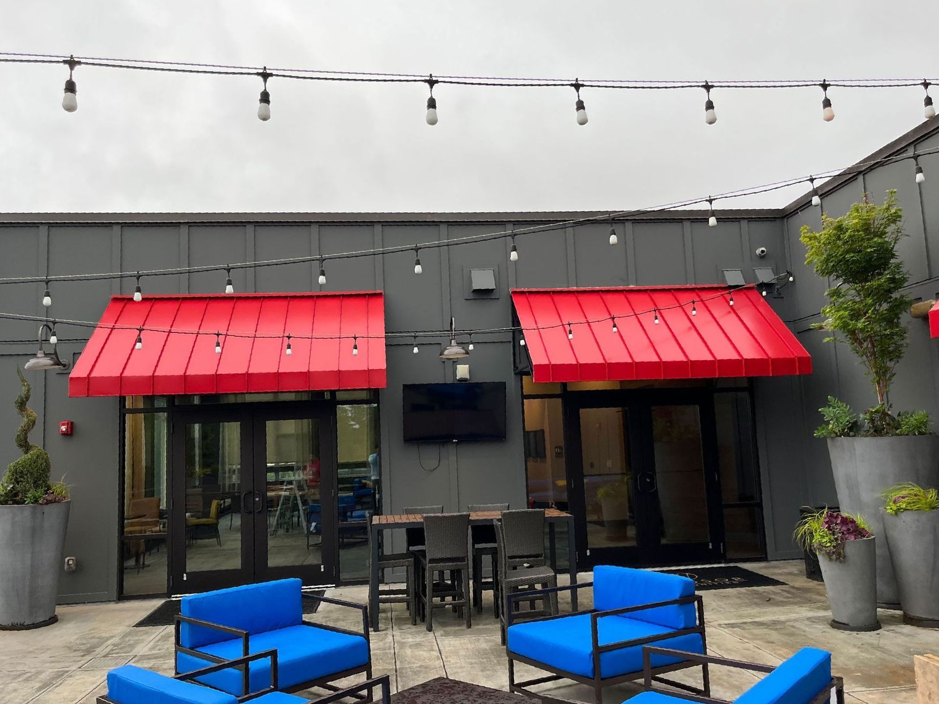 Rooftop patio with red awnings, string lights, blue seating, and gray building exterior.