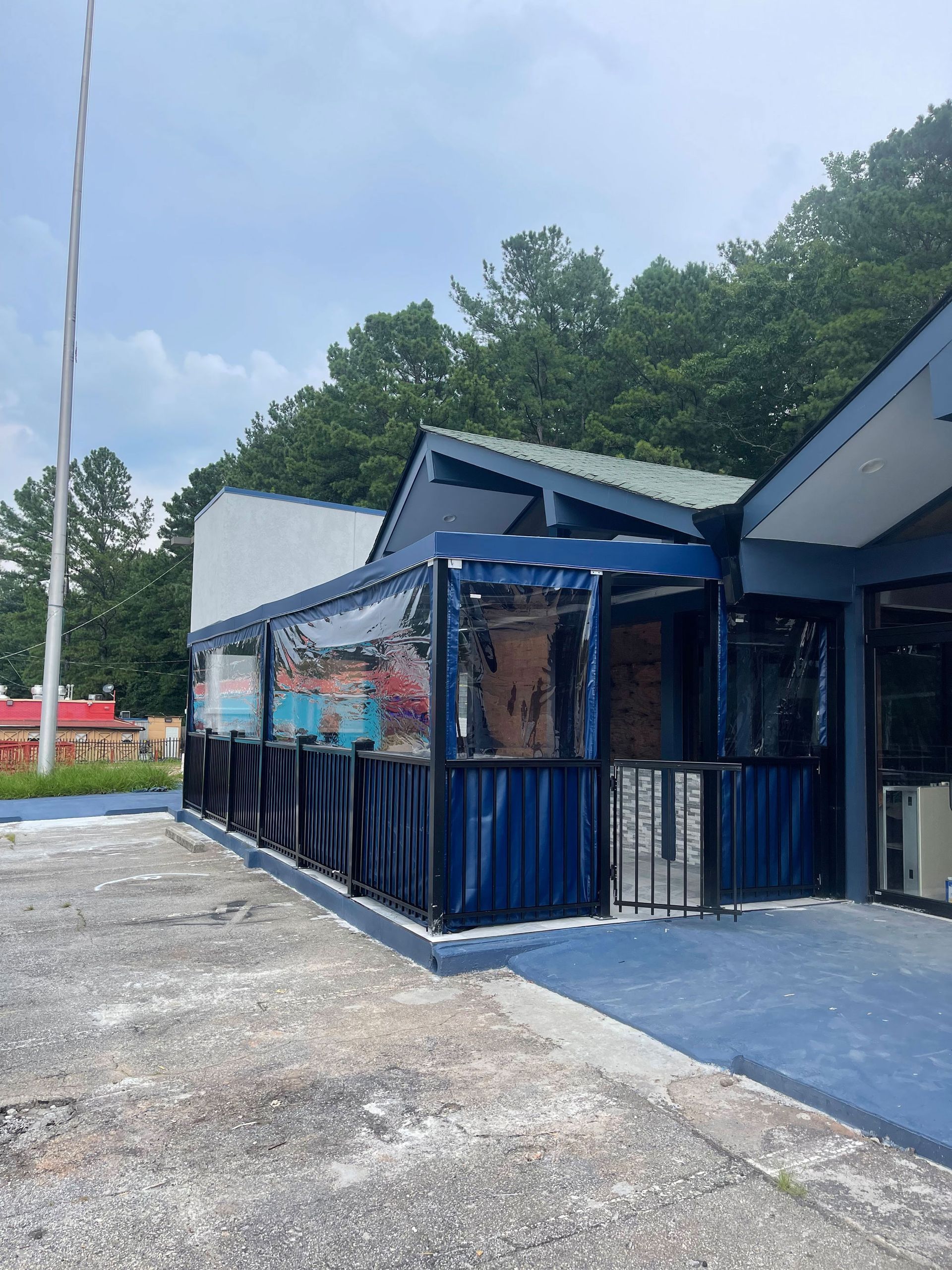 Restaurant exterior with blue trim, covered patio, clear plastic sides. Trees in the background, blue sky.