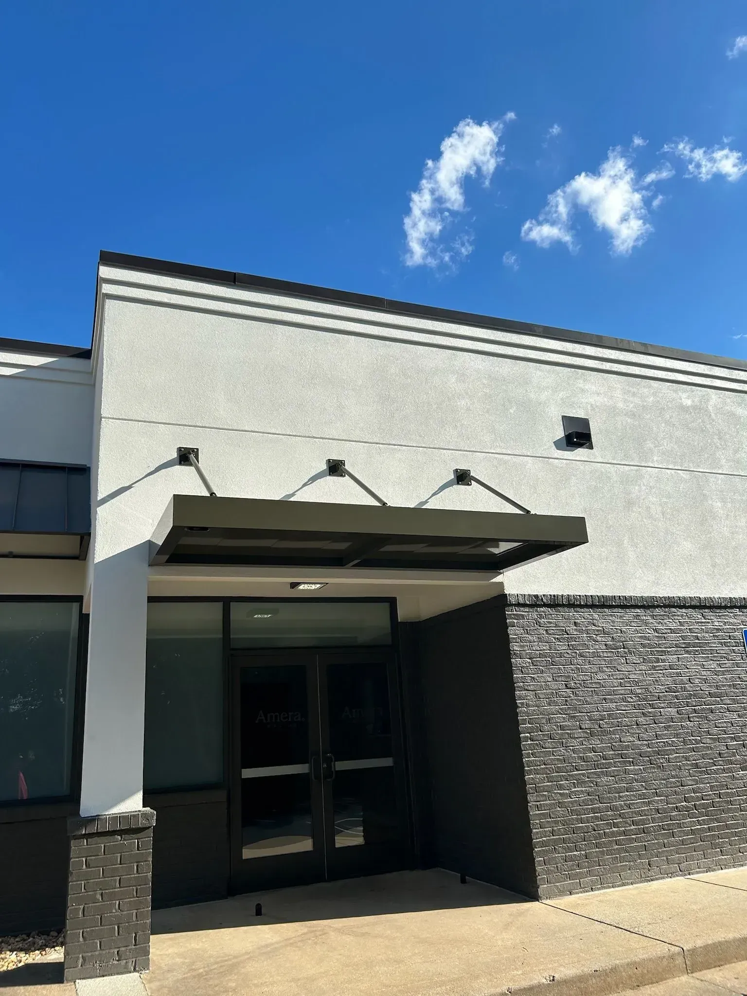 Gray commercial building exterior with black awning and doors under a blue sky.