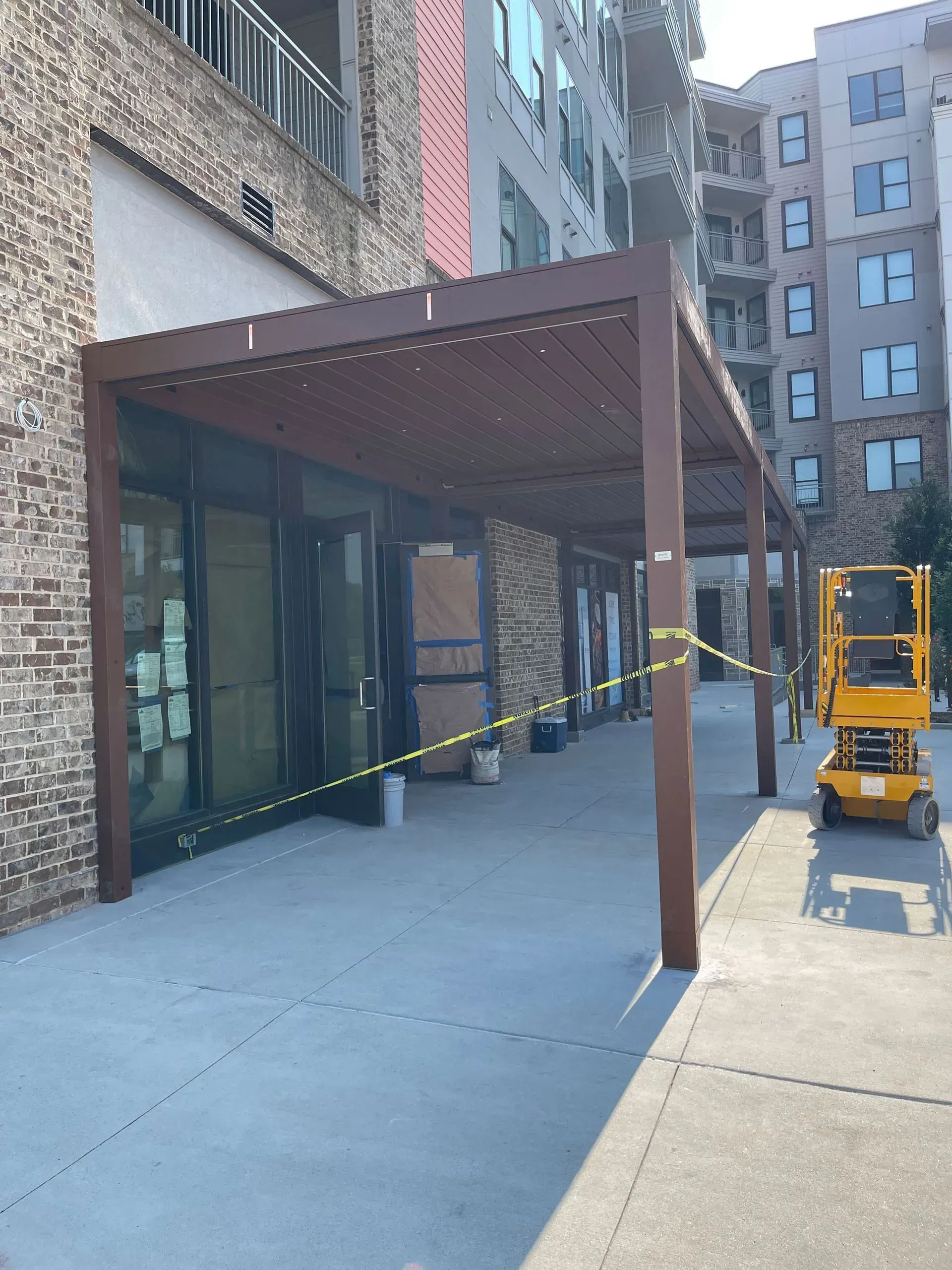 Pergola shading storefronts along a sidewalk, next to a multi-story brick building. A yellow lift sits on the right.