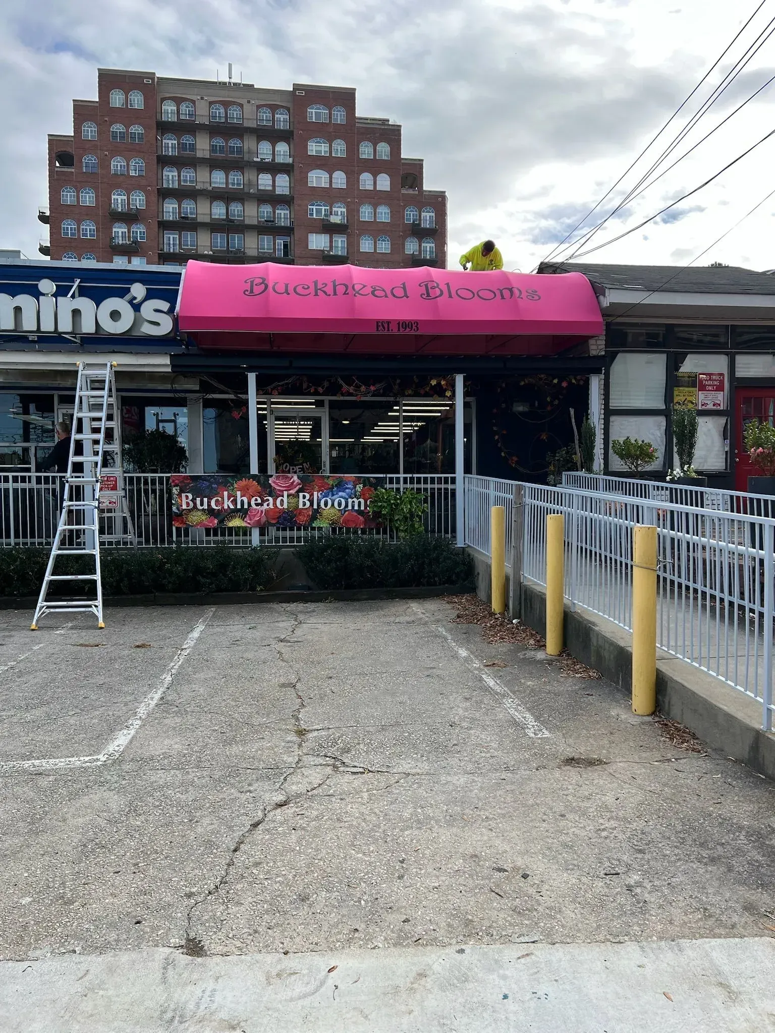 Storefront with pink awning: "Flowers on Main". Next to Domino's, tall brick building in background. Cloudy day.