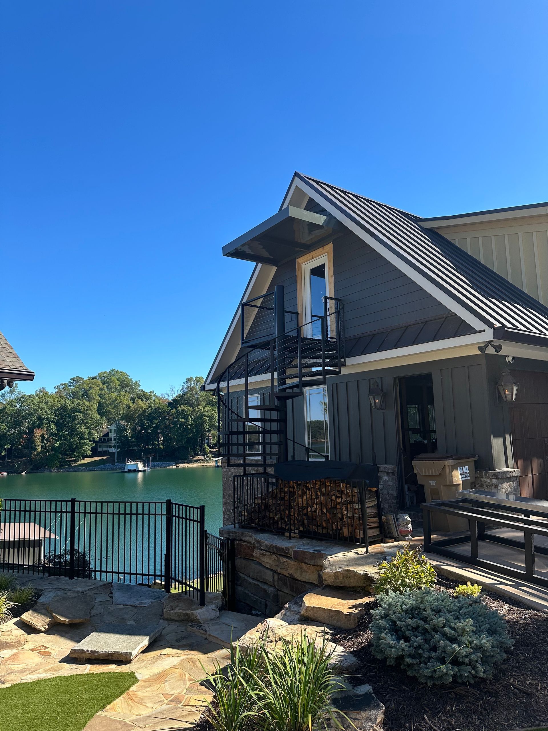 Lakeside house with spiral staircase, dark siding, and a black metal fence on a sunny day.