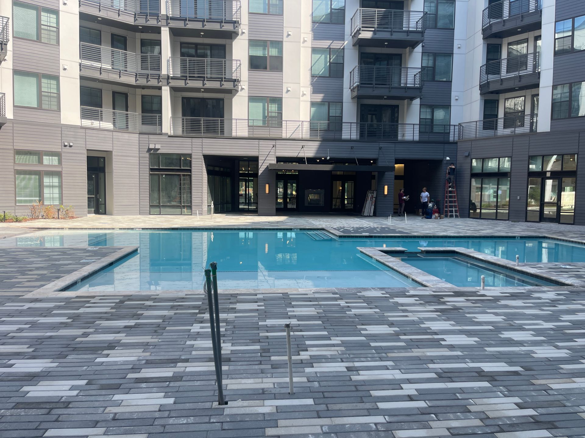Pool in front of multi-story building, blue water, gray brick patio, with a dark entrance and balconies.