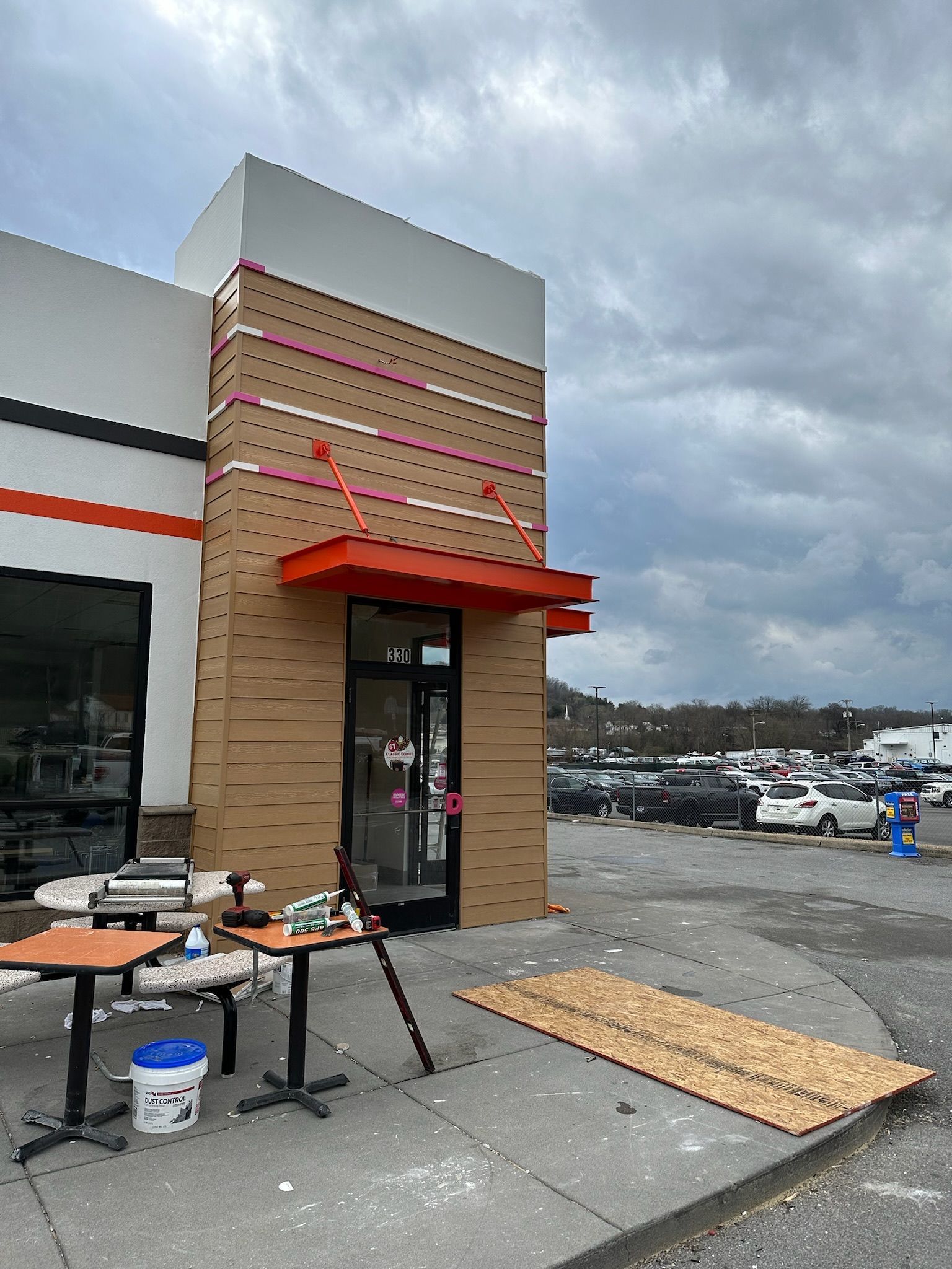 Exterior of a building under construction. Brown siding, orange accents, cloudy sky. Construction materials and tables present.