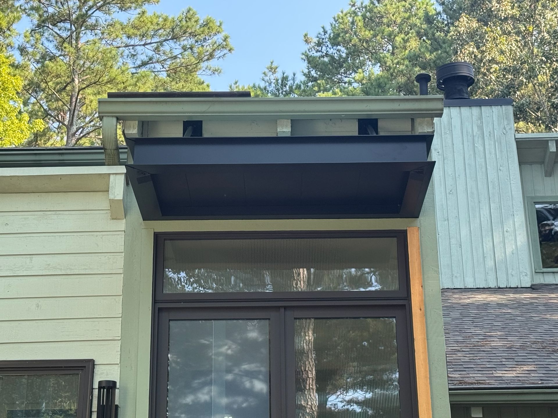 Black awning above a window on a light-colored building with a dark brown frame, set against a backdrop of trees.