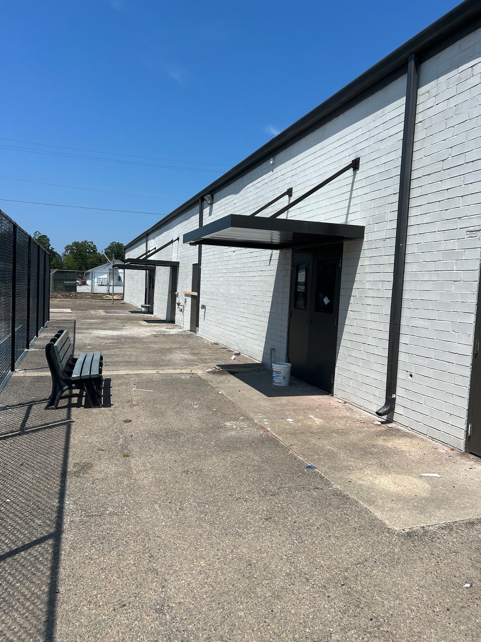 Exterior of a building with multiple doors, each with a small awning. A bench sits in front of a chain-link fence.