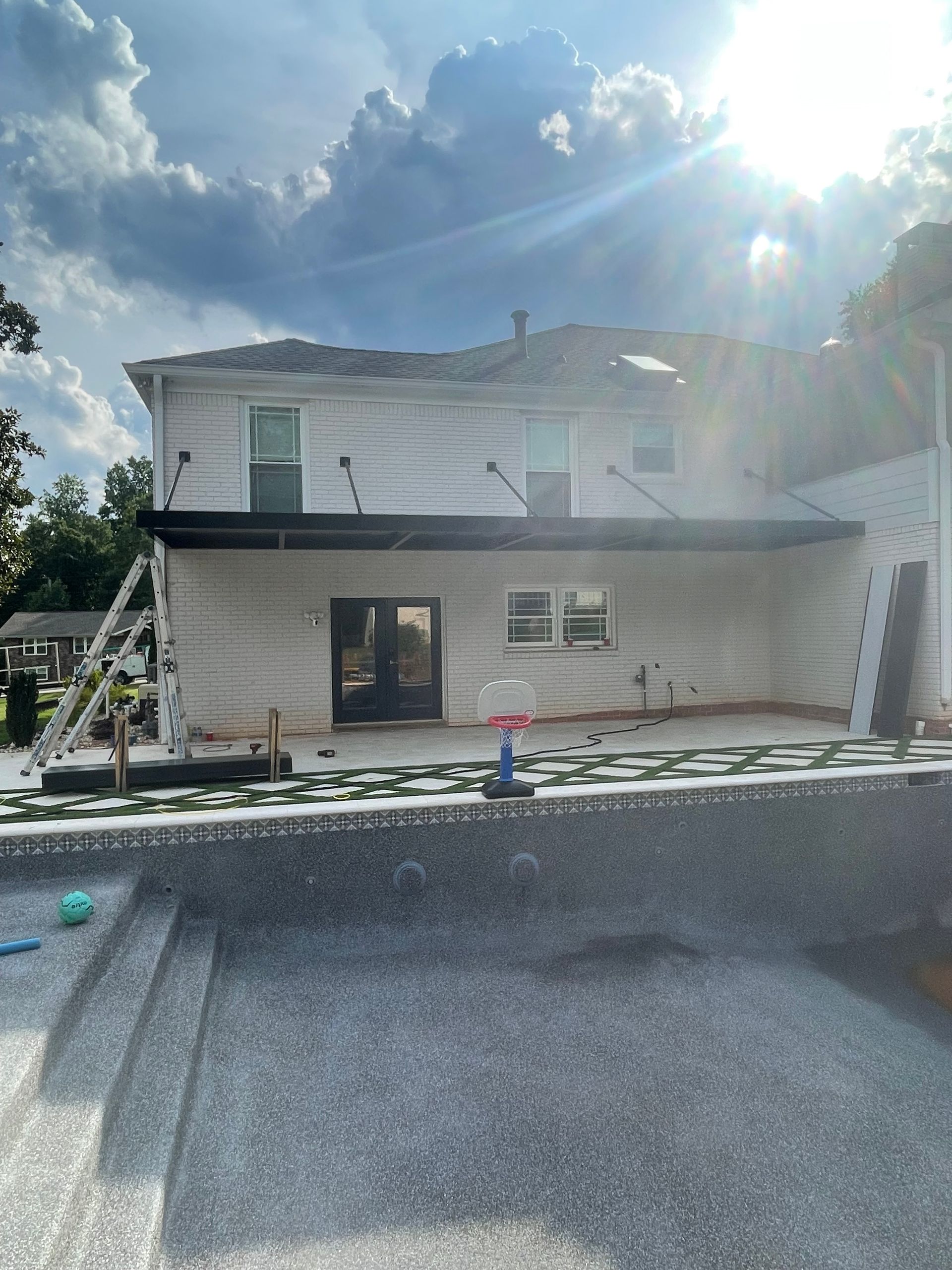 Back of a house with a pool under construction. Stone pool, patio with supports, cloudy sky.