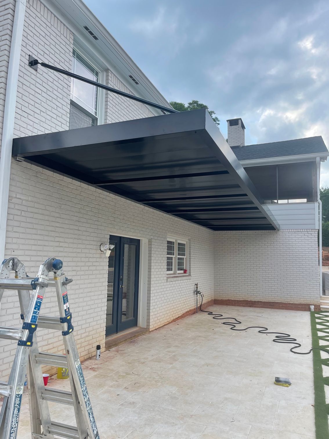 Black awning attached to a white brick house, over patio area. Ladder in the foreground.