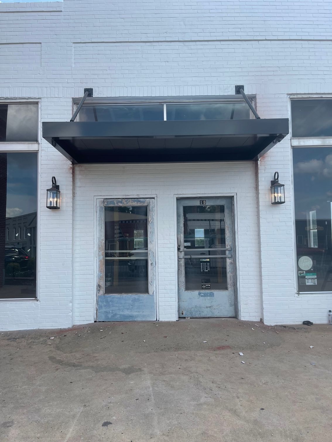 White brick building with two glass doors under a black awning. Exterior lamps flank the doors.