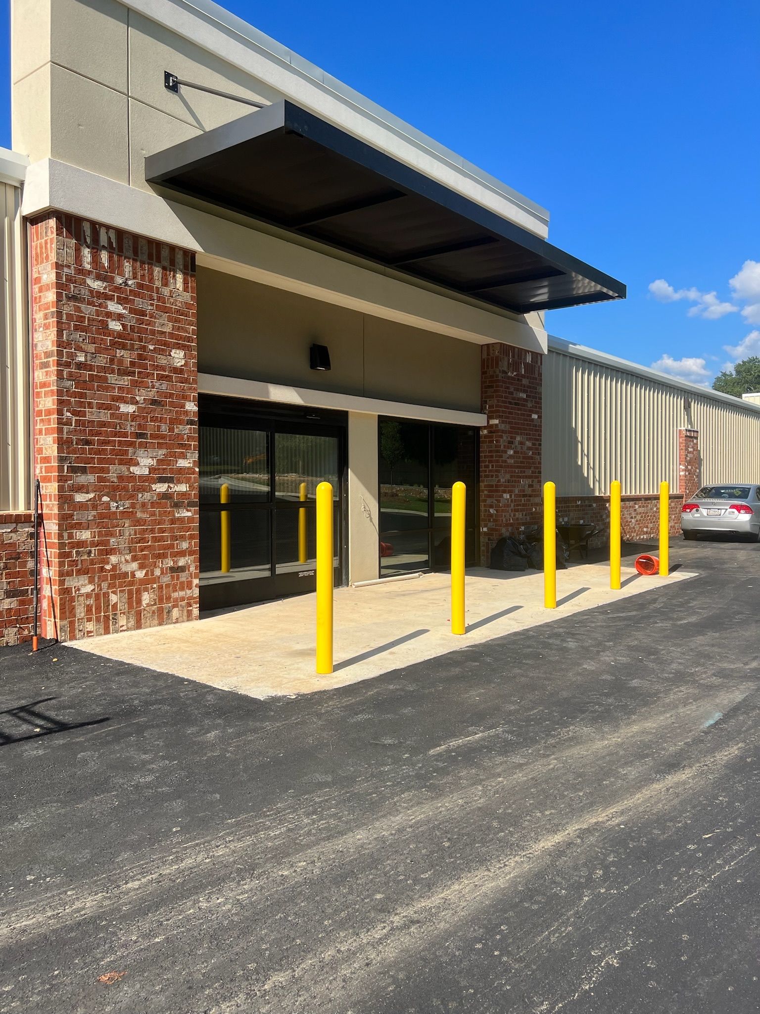 Exterior of a building with brick accents, automatic doors, a black awning, and yellow bollards.