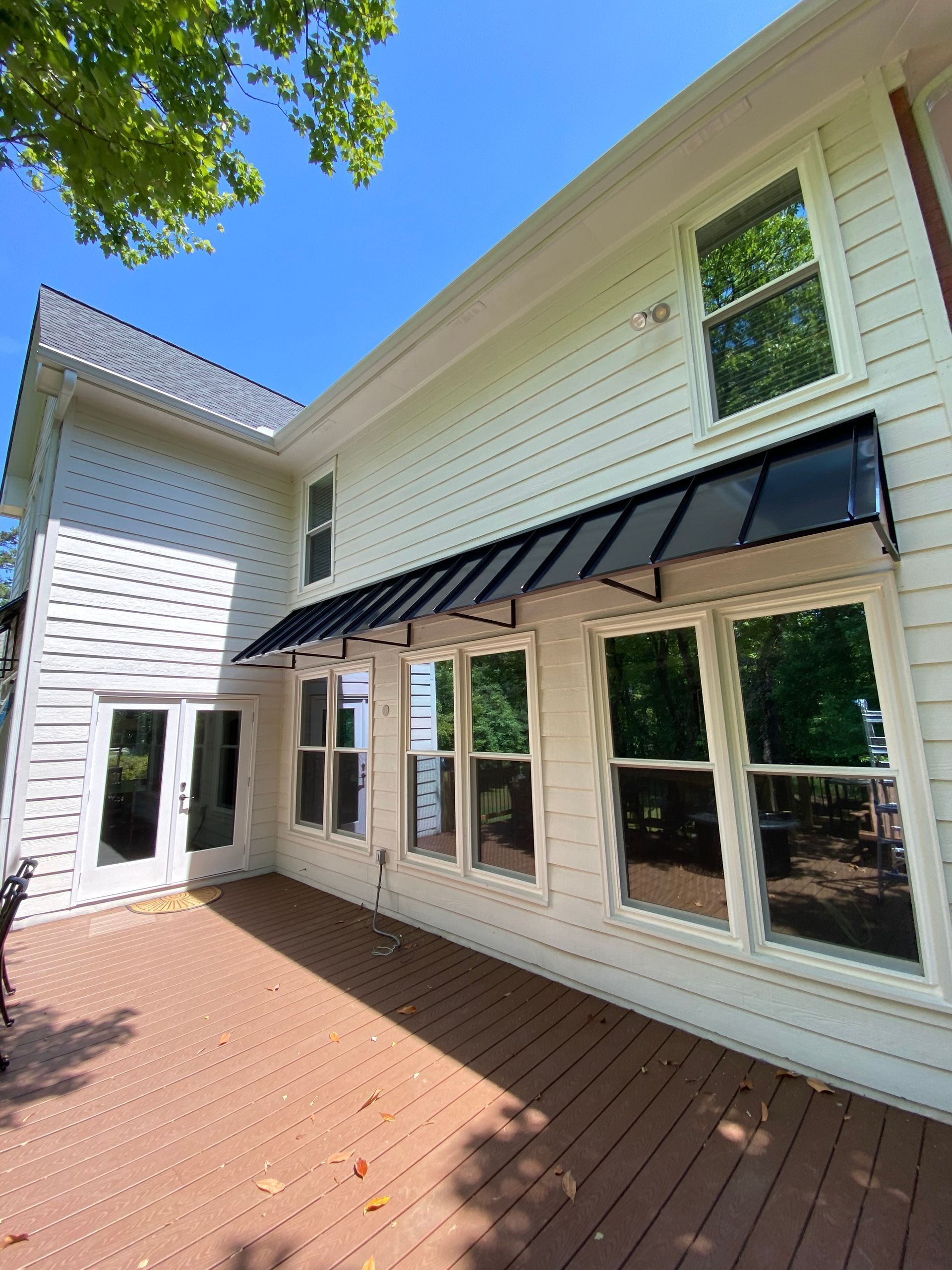 Exterior view of a white house with a black awning over windows on a wooden deck. Sunny day.