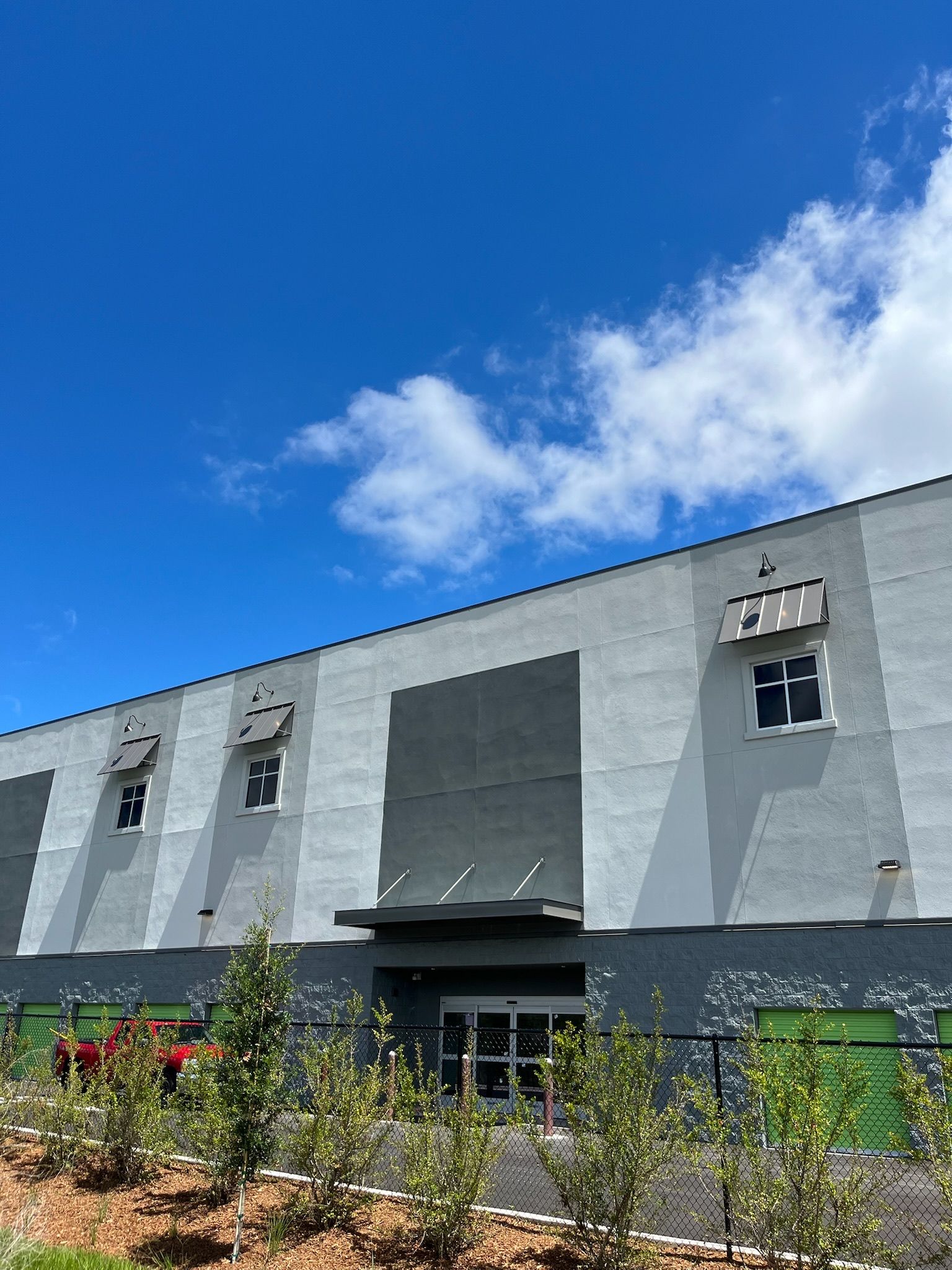 Gray and white building under blue sky with clouds; bushes and a fence in front.