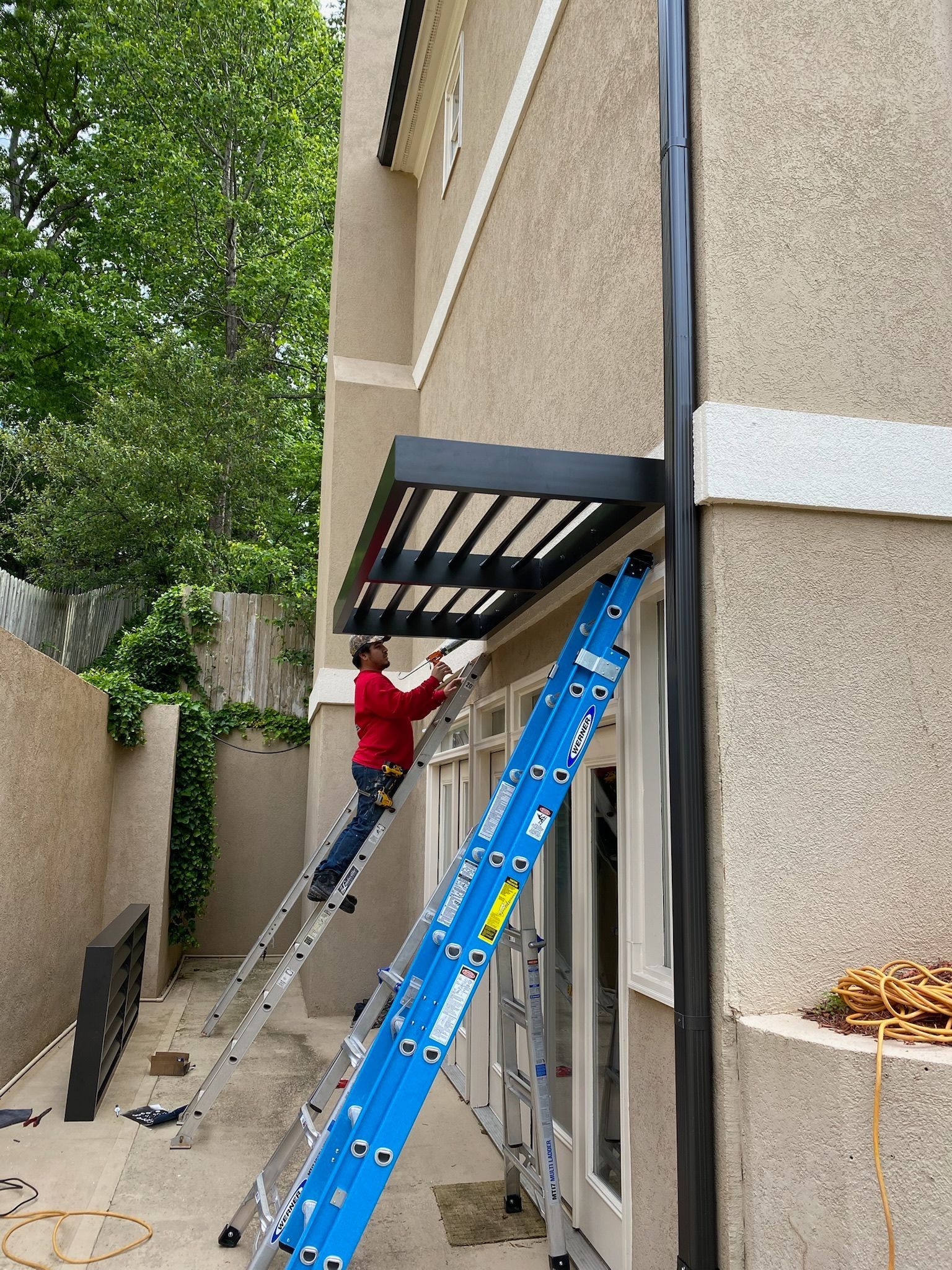Person on ladder installing awning over glass doors of a tan building.