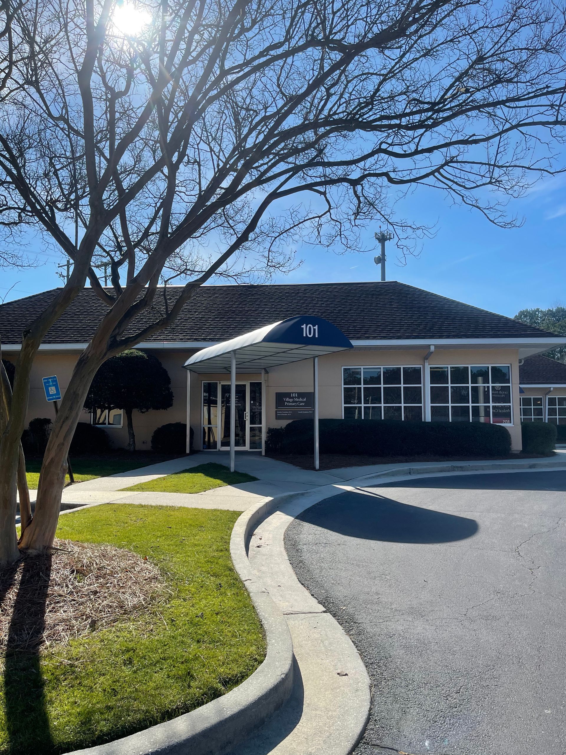 Tan building with a dark tiled roof and a small awning, viewed from a tree-lined driveway.
