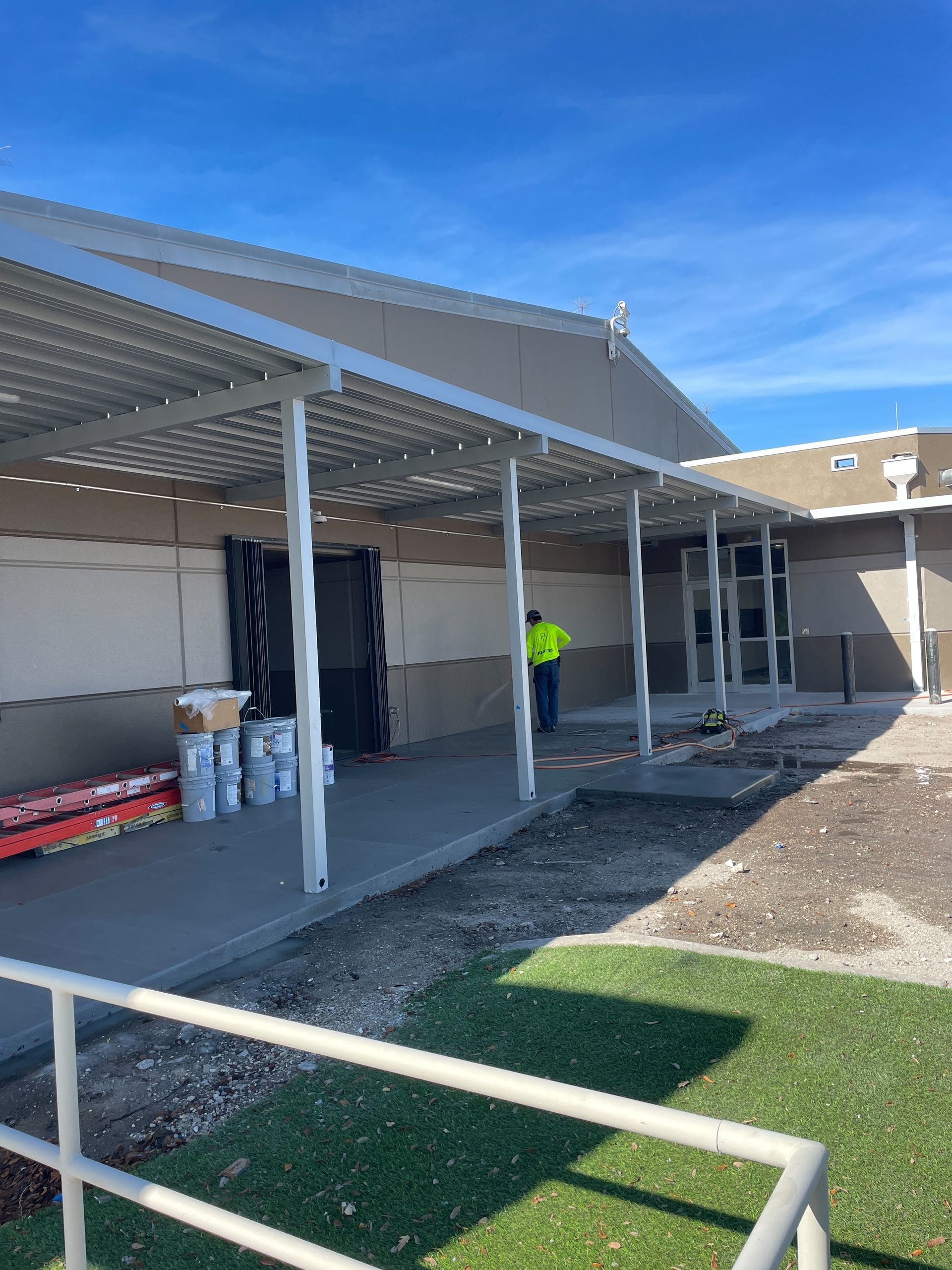 Construction site: a covered walkway with a worker, concrete work, and supplies against a building under a blue sky.