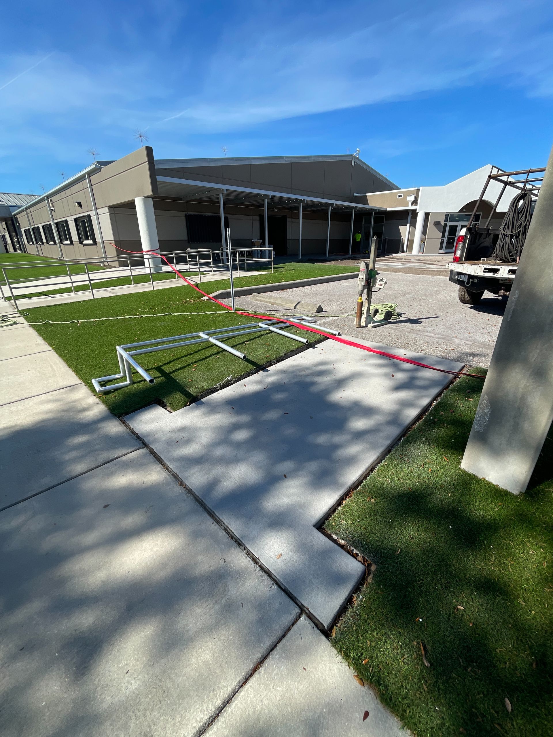 Construction site with building, grass, concrete, and equipment under a blue sky.