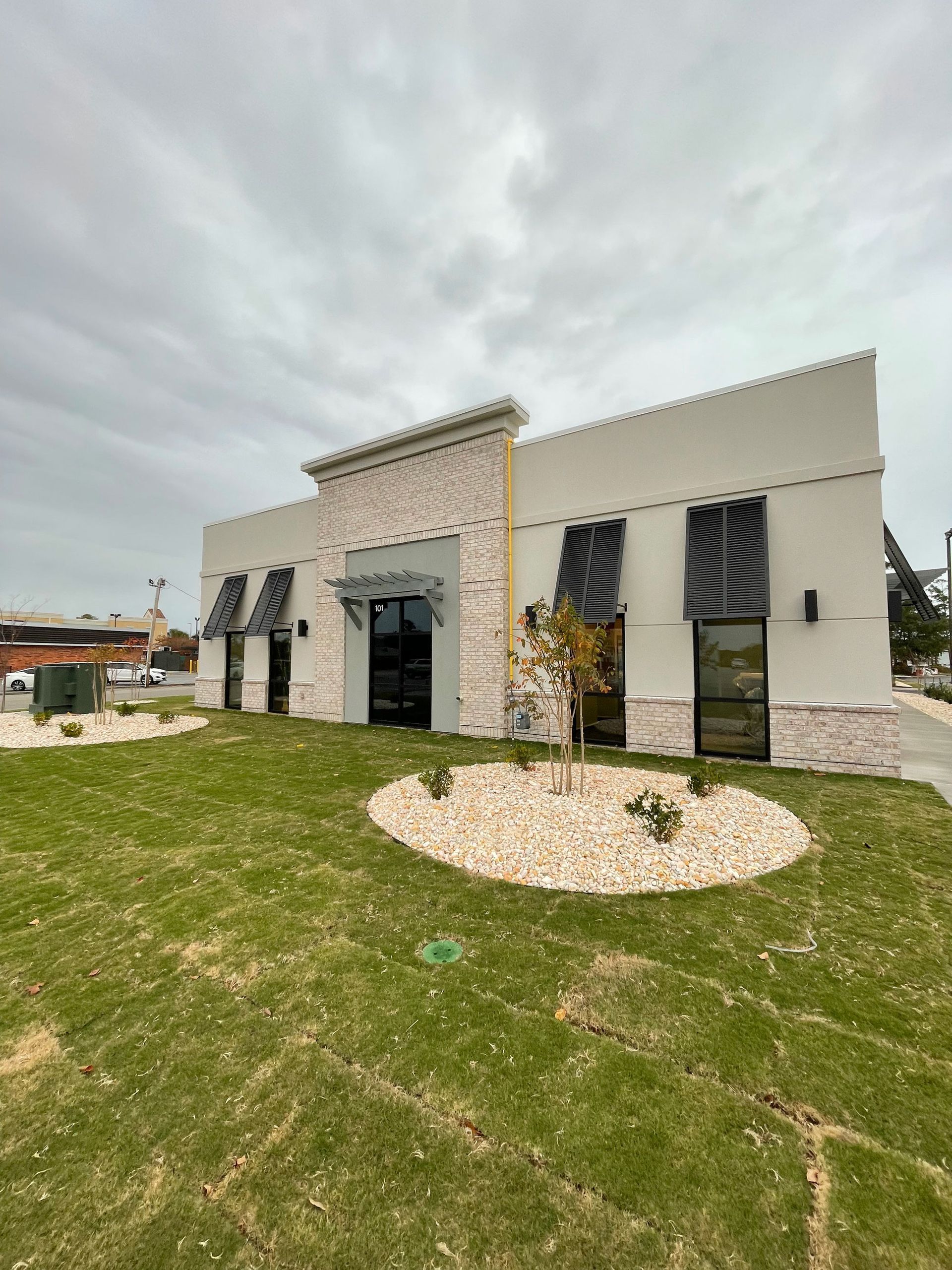 Building with light beige walls, black window coverings, and a stone facade under an overcast sky.