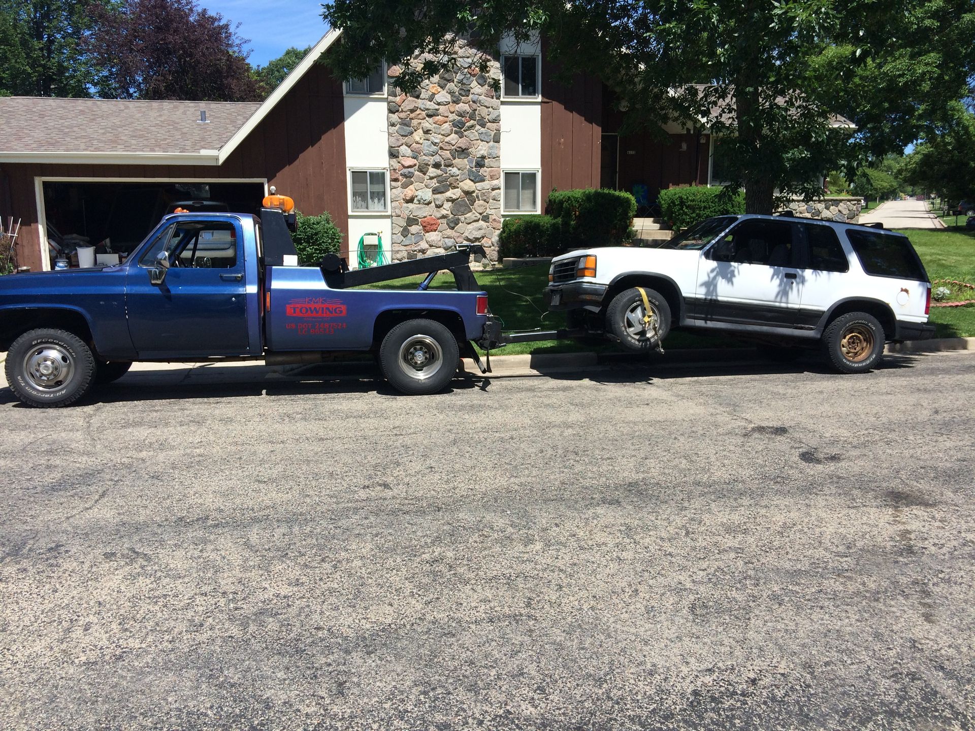 A blue tow truck is towing a white suv in front of a house.