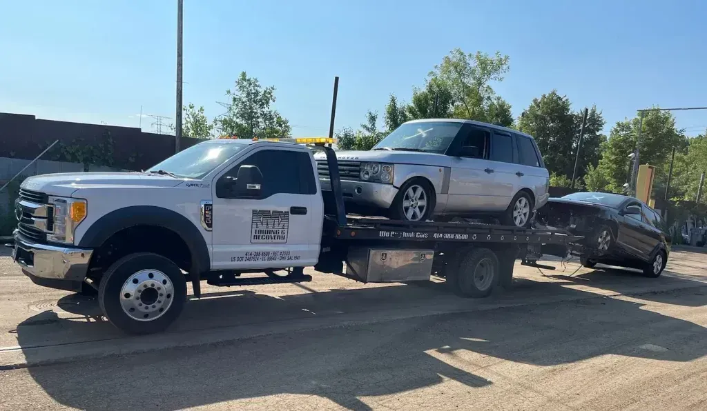 A tow truck is towing two cars in a parking lot.