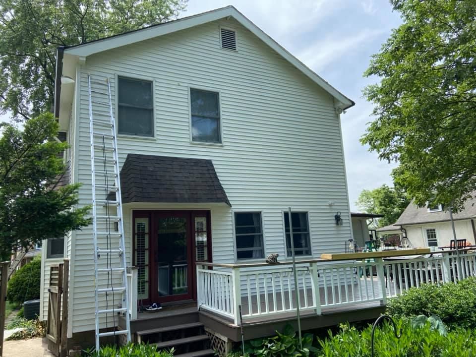 Back of a two-story white house with a deck, dark roof, windows, and ladder leaning against the side.