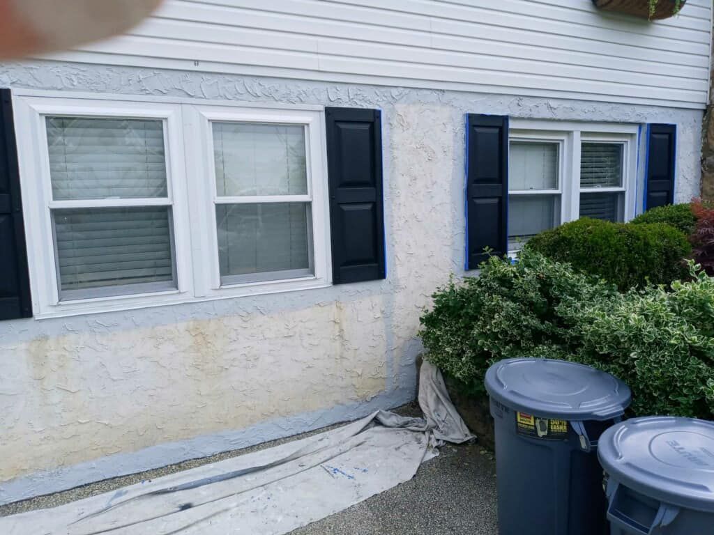 Two white-framed windows with black shutters on a stucco house. Trash cans and bushes are in front.