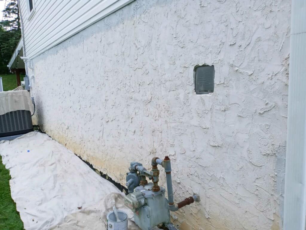 Exterior wall of a house with stucco finish; gas meter at bottom; protective covering on ground.