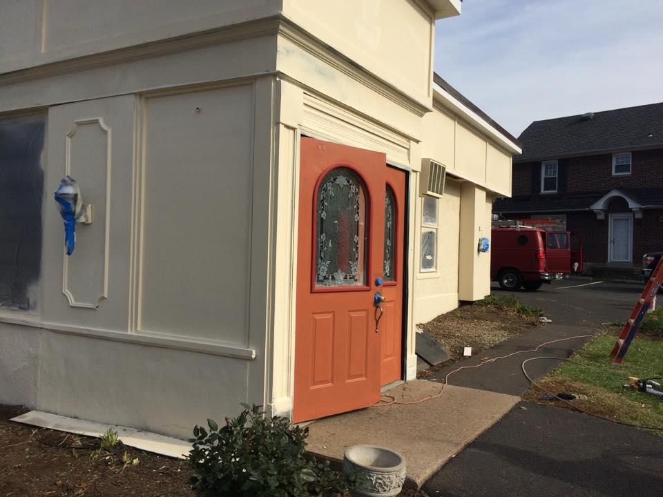 Exterior of a building with an open orange door, cream walls, and a small concrete walkway.