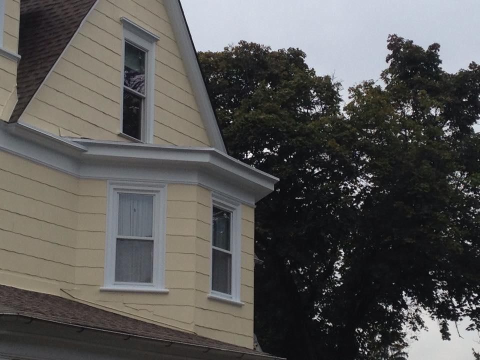 Yellow house with white trim, bay window, and a tall tree in the background. Overcast sky.