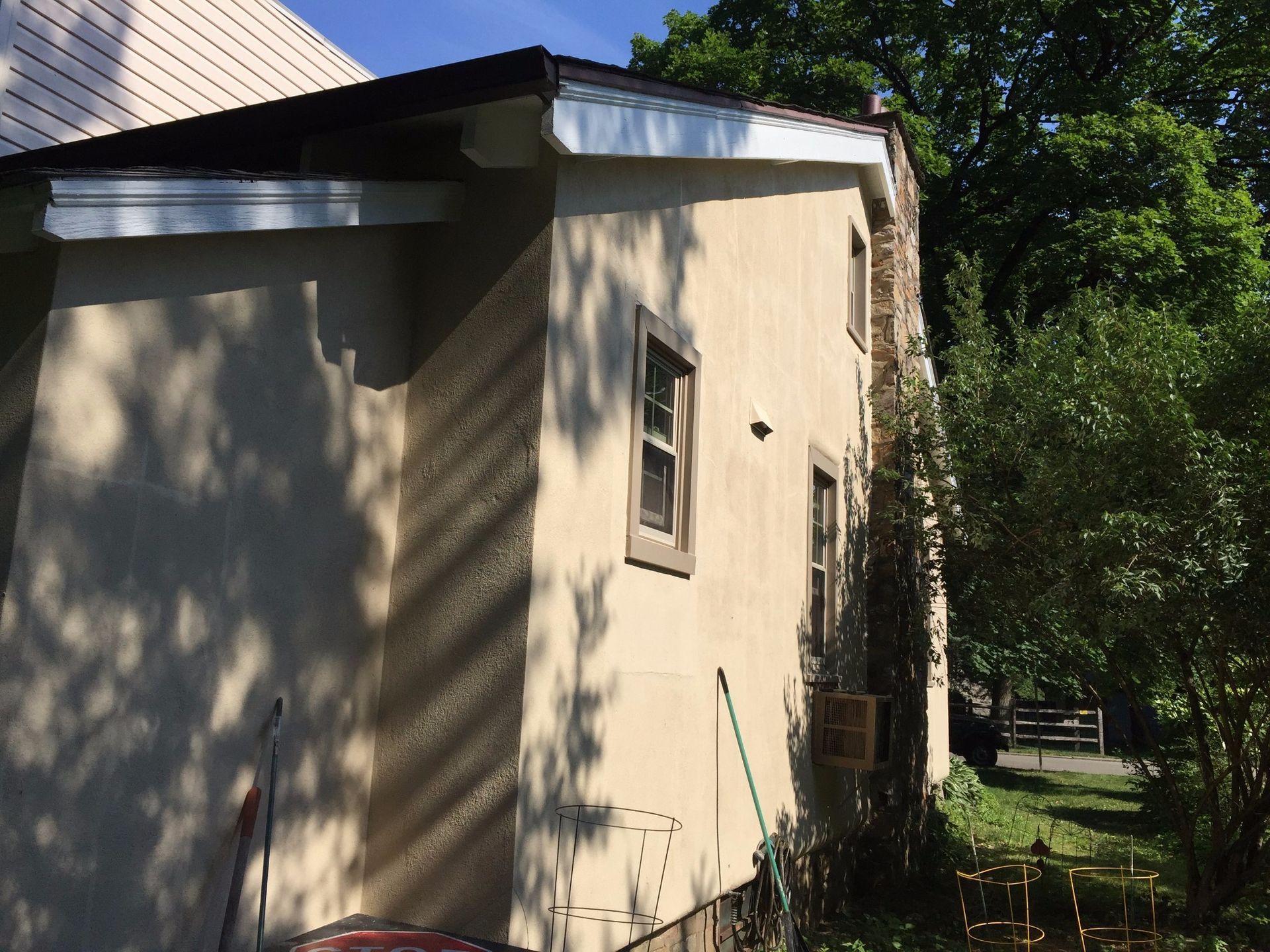 Beige house exterior with dark roof, two windows, and chimney next to foliage.
