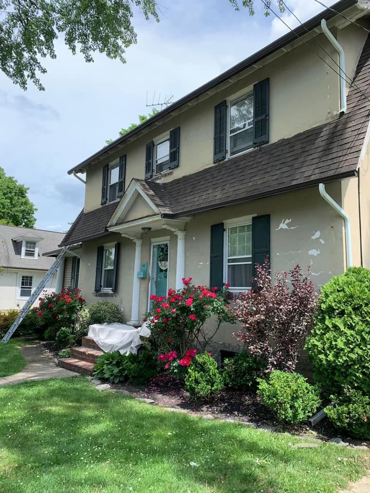 Two-story beige house with dark roof and green shutters, red flowers and shrubs in front yard.