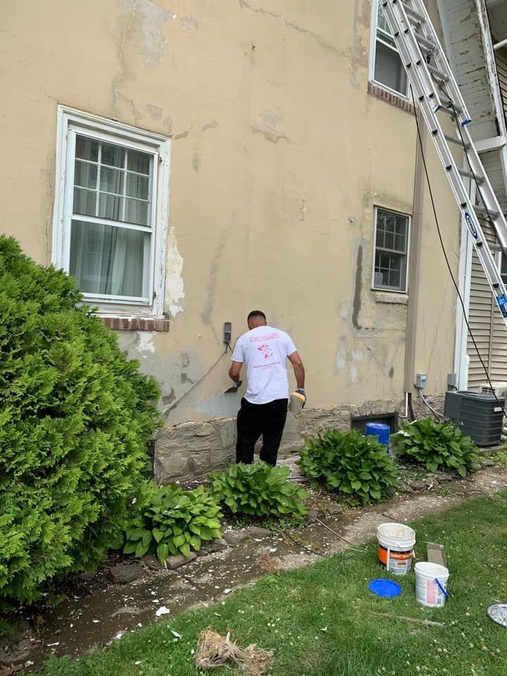 Man scraping stucco on a building exterior. A ladder is propped against the side, surrounded by landscaping and tools.