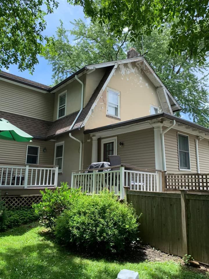 Beige two-story house with brown roof, white porch and railing, surrounded by green trees and bushes.