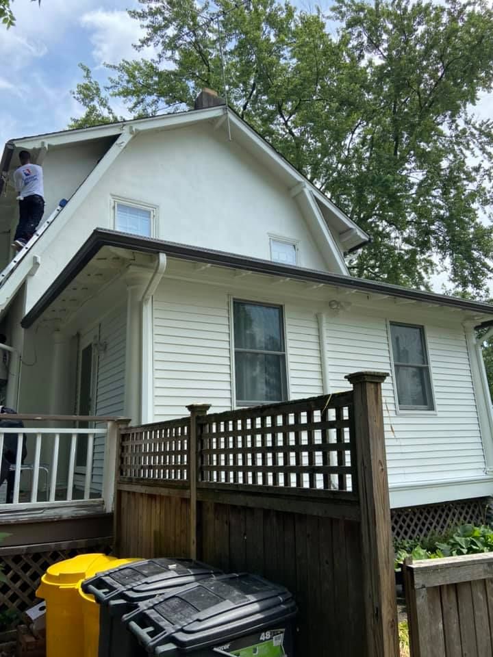 Person on a ladder painting a white house with a dark roof trim and a wooden fence.