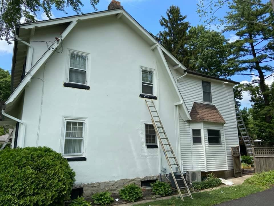 White house exterior with ladders, undergoing painting. Sunny day, greenery in background.