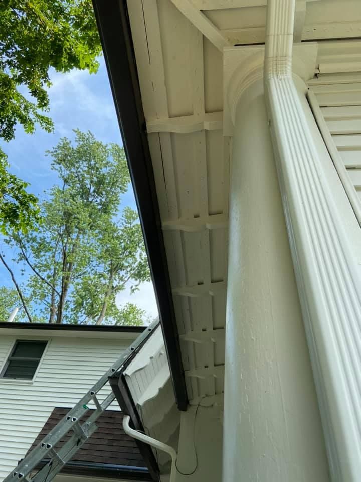 Black gutter along white house trim, ladder, and trees against a blue sky.