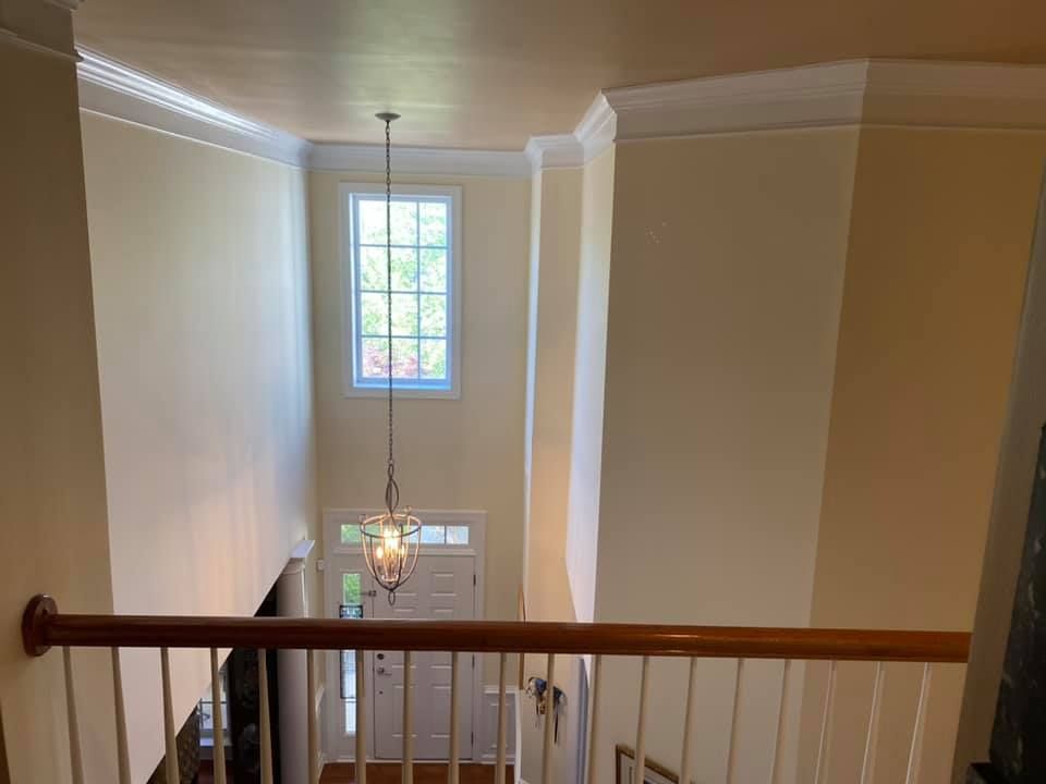 View from a second-floor landing looking down at a front door, chandelier, and tall window. Cream walls, wood railing.